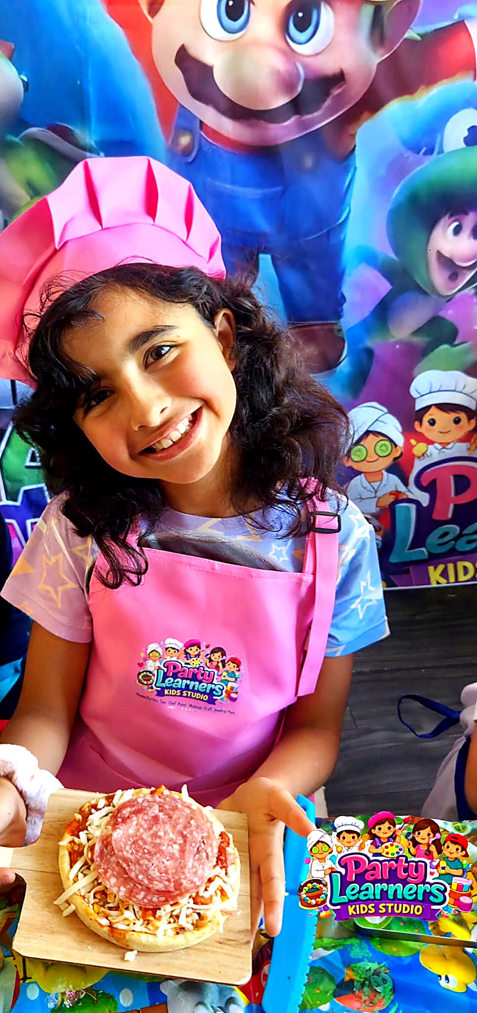 Girl in pink chef hat and Party Learners Kids Studio apron proudly showing her decorated mini pizza at a Chef Party — Decorating Experience