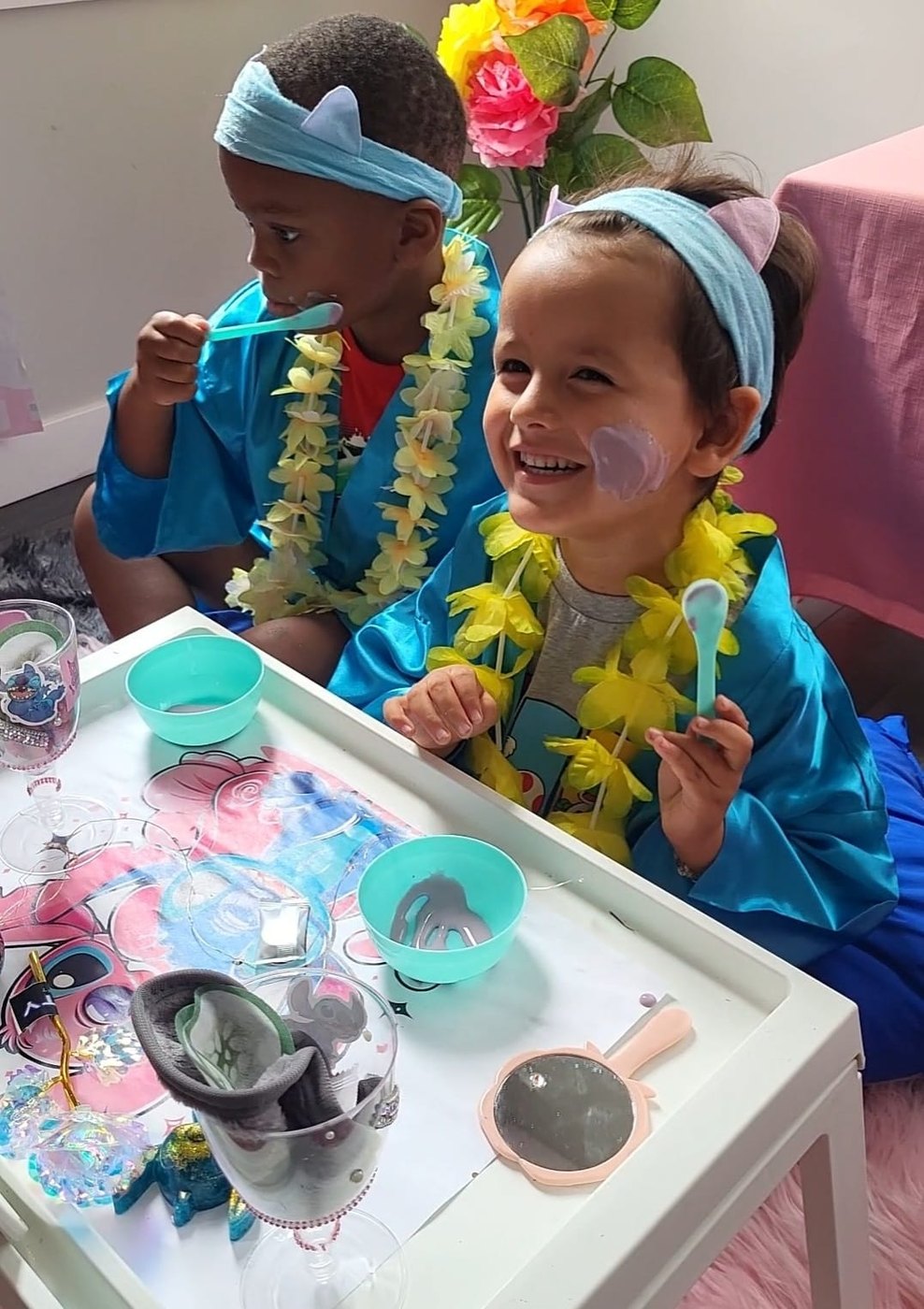 A boy and a girl in blue robes and floral leis enjoying spa craft activities at a Divas Glam Spa Party