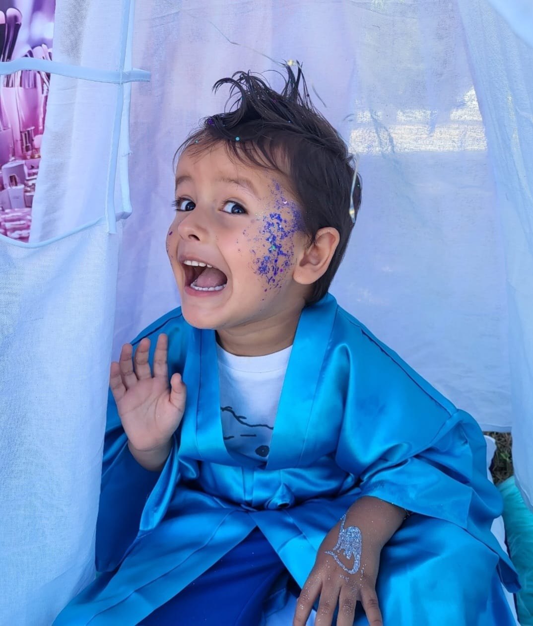 Joyful boy in a blue silky robe with sparkle on his cheek at a Divas Glam Spa Party — boys are welcome too