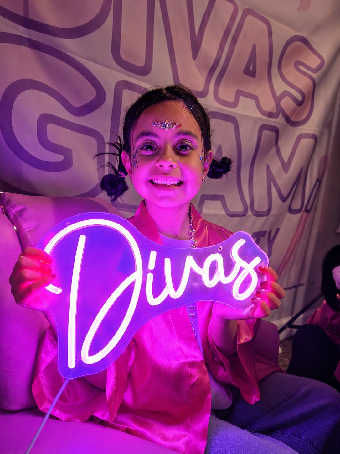 Birthday girl with sparkle face gems holding the glowing pink Divas neon sign in front of the Divas Glam backdrop at a Divas Glam Spa Party