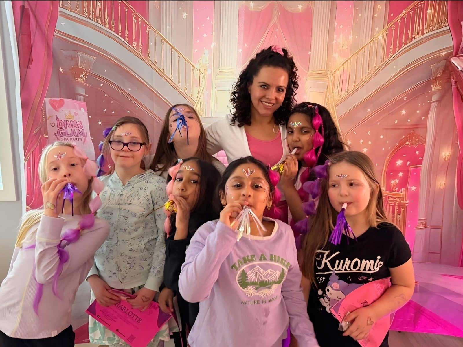 Group of girls with face gems, sparkle hair extensions and party blowers at a Divas Glam Spa Party with Party Learners host Jenn in front of a pink castle backdrop