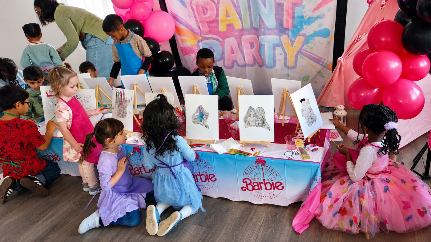 Kids painting at wooden easels at a Barbie-inspired Paint Boho Party + Craft with PAINT PARTY backdrop, pink balloons and Barbie tablecloth at Party Learners Kids Studio