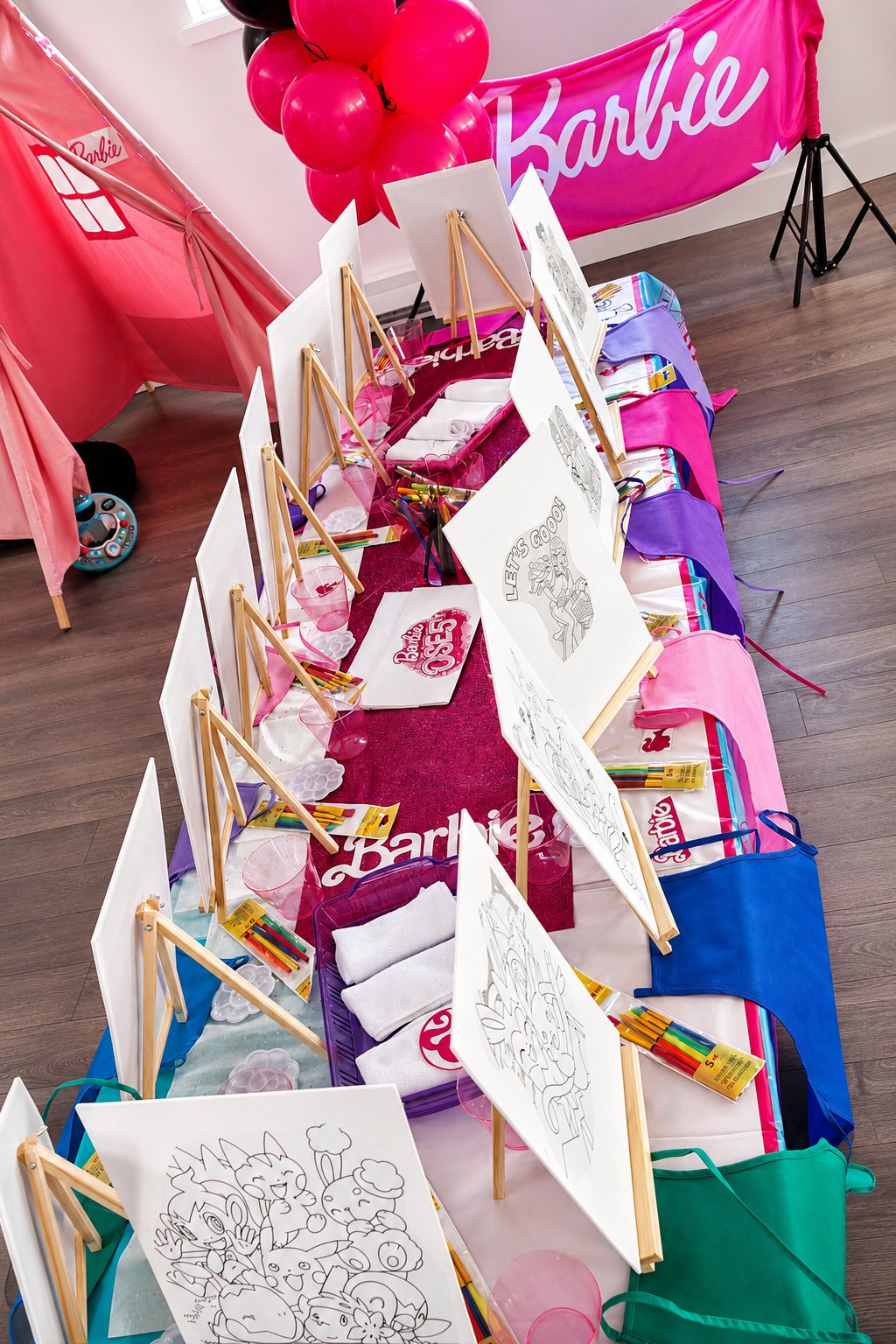 Wide top-down view of a Paint Boho Party + Craft long Barbie-themed table with multiple wooden easels, blank canvases, paint kits and pink Barbie tipi at a Party Learners Kids Studio party